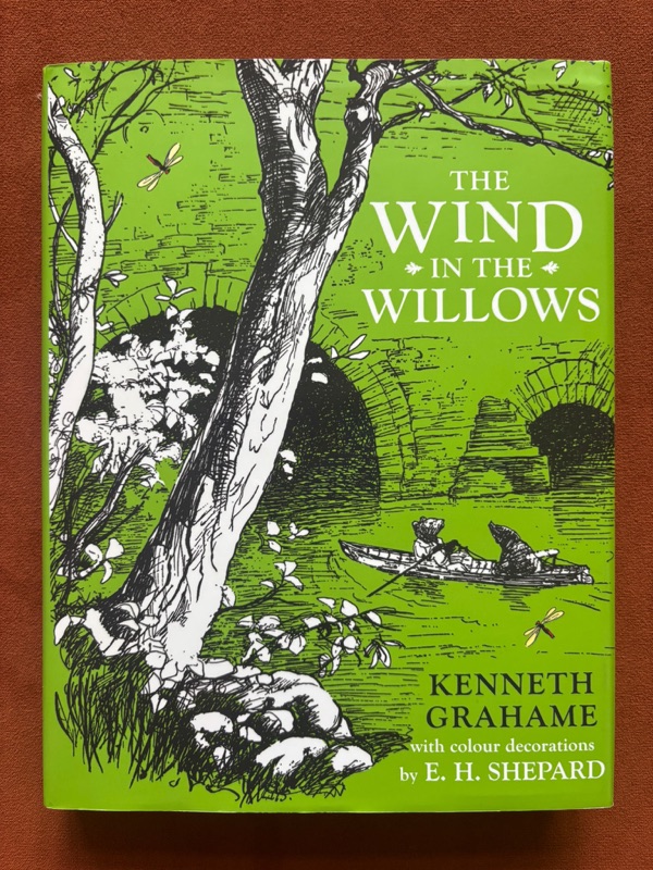 Robbie's copy of The Wind in the Willows by Kenneth Grahame. A green clothbound hardcover edition with cover illustrations by E. H. Shepard showing a tree silhouette over a riverbank, with two small figures in a rowing boat below.